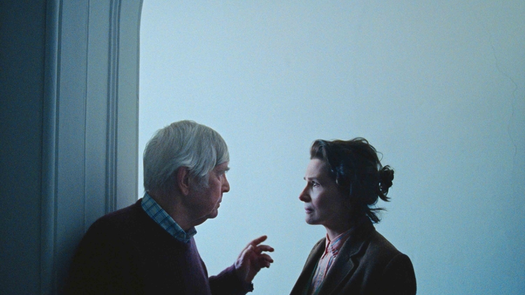 Two people in profile face each other in a dimly lit room with a plain light blue wall in the background.