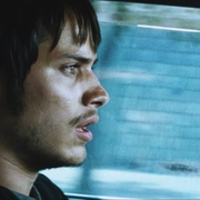 Side profile of a young man driving a car, looking intently forward with a blurred blue background outside the window.