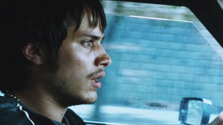 Side profile of a young man driving a car, looking intently forward with a blurred blue background outside the window.