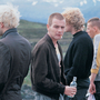Four young men standing outdoors near train tracks, one smoking a cigarette and holding a bottle, another holding a can, all dressed casually.