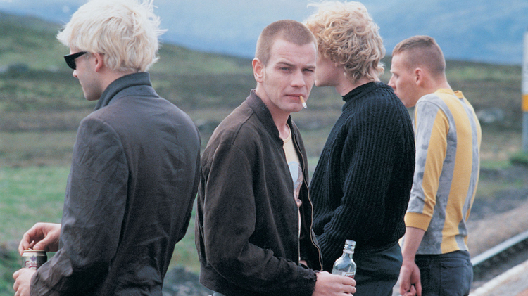 Four young men standing outdoors near train tracks, one smoking a cigarette and holding a bottle, another holding a can, all dressed casually.