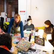Diverse group creates art around a craft table in a bright studio, with paints, collage materials, colourful projects, and a mural behind them.