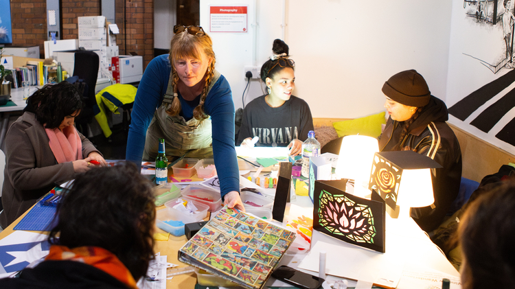 Diverse group creates art around a craft table in a bright studio, with paints, collage materials, colourful projects, and a mural behind them.