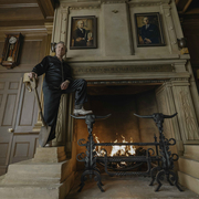 Man in black pyjamas stands beside a roaring fireplace in a wood-panelled room displaying portraits, an antique clock, and mounted antlers.