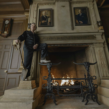Man in black pyjamas stands beside a roaring fireplace in a wood-panelled room displaying portraits, an antique clock, and mounted antlers.