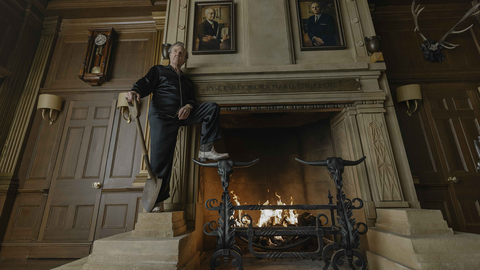 Man in black pyjamas stands beside a roaring fireplace in a wood-panelled room displaying portraits, an antique clock, and mounted antlers.