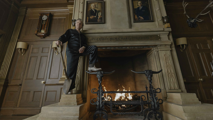 Man in black pyjamas stands beside a roaring fireplace in a wood-panelled room displaying portraits, an antique clock, and mounted antlers.