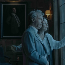 A man and woman look out of a large window. The man looks confident, but concerned and the woman looks anxious. The room is dimly lit and a portrait of a man in a business suit hangs in the background.