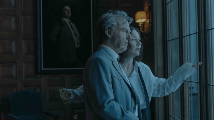 A man and woman look out of a large window. The man looks confident, but concerned and the woman looks anxious. The room is dimly lit and a portrait of a man in a business suit hangs in the background.