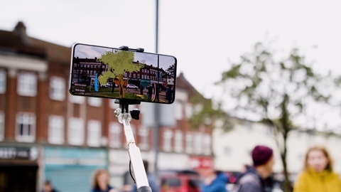 Smartphone on a selfie stick displays an augmented-reality view of a historic street scene.