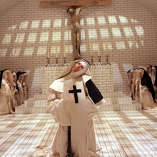 Nuns in white and black habits gathered in a tiled chapel, one nun in front with a large cross looks upward beneath a crucifix.