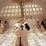 Nuns in white and black habits gathered in a tiled chapel, one nun in front with a large cross looks upward beneath a crucifix.