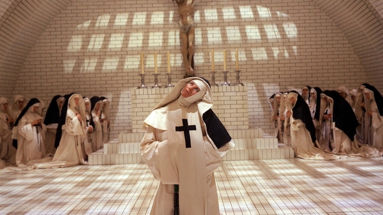 Nuns in white and black habits gathered in a tiled chapel, one nun in front with a large cross looks upward beneath a crucifix.
