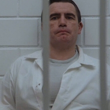 Man in a white prison uniform sits behind bars, facing a blurred figure in the foreground.