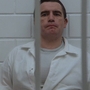 Man in a white prison uniform sits behind bars, facing a blurred figure in the foreground.