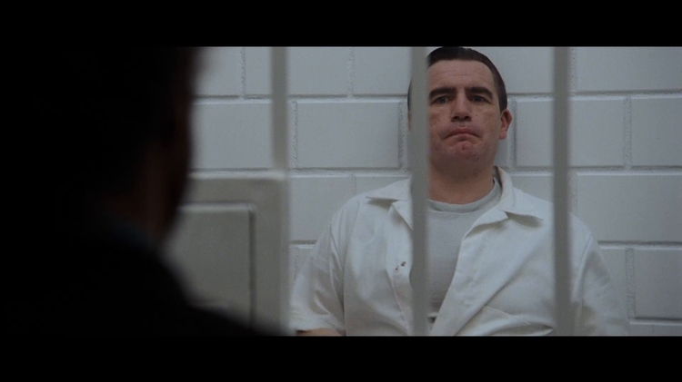 Man in a white prison uniform sits behind bars, facing a blurred figure in the foreground.