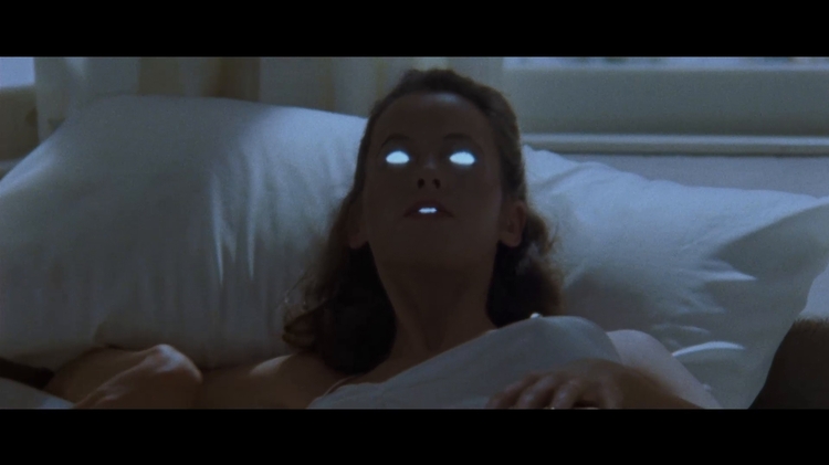 A person lies in bed in darkness, their eyes and mouth glowing unnaturally white.