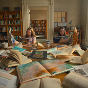 Two girls reading, surrounded by open books and maps.