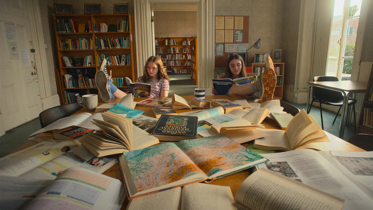 Two girls reading, surrounded by open books and maps.