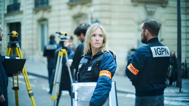 Police investigators in a city street, with a blonde woman facing the camera and surveying equipment in the foreground.