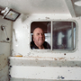 Bald man looks through a small sliding window in the worn white cabin of a boat.