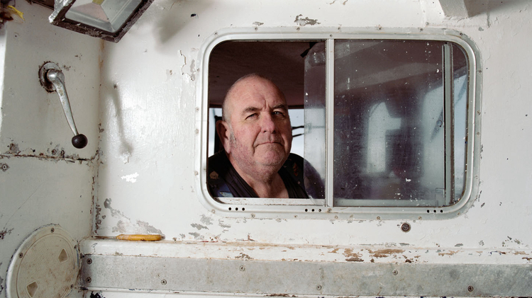 Bald man looks through a small sliding window in the worn white cabin of a boat.