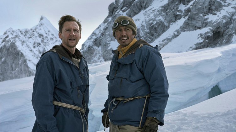 Two men in winter clothing stand on a snowy mountain, smiling with climbing ropes and gear.